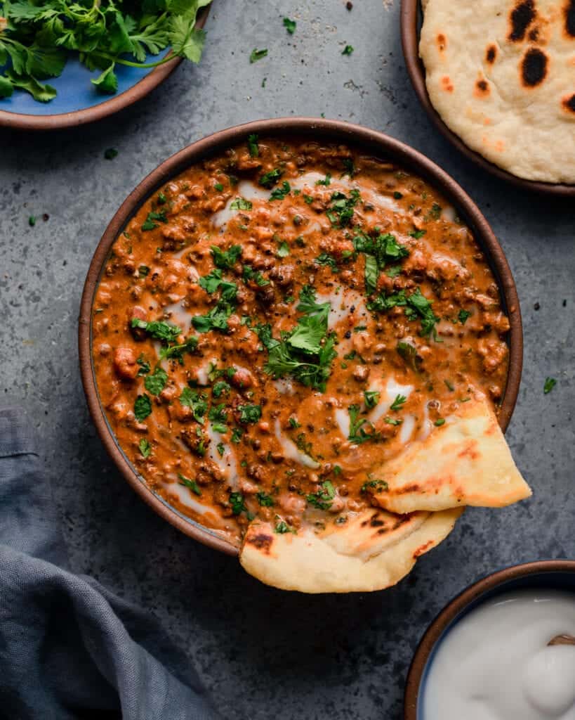 bowl of creamy vegan dal makhani with naan, yogurt, and cilantro on blue background