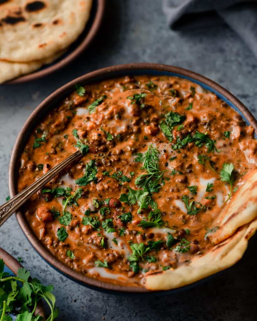 closeup bowl of creamy vegan dal makhani with spoon and naan on blue background