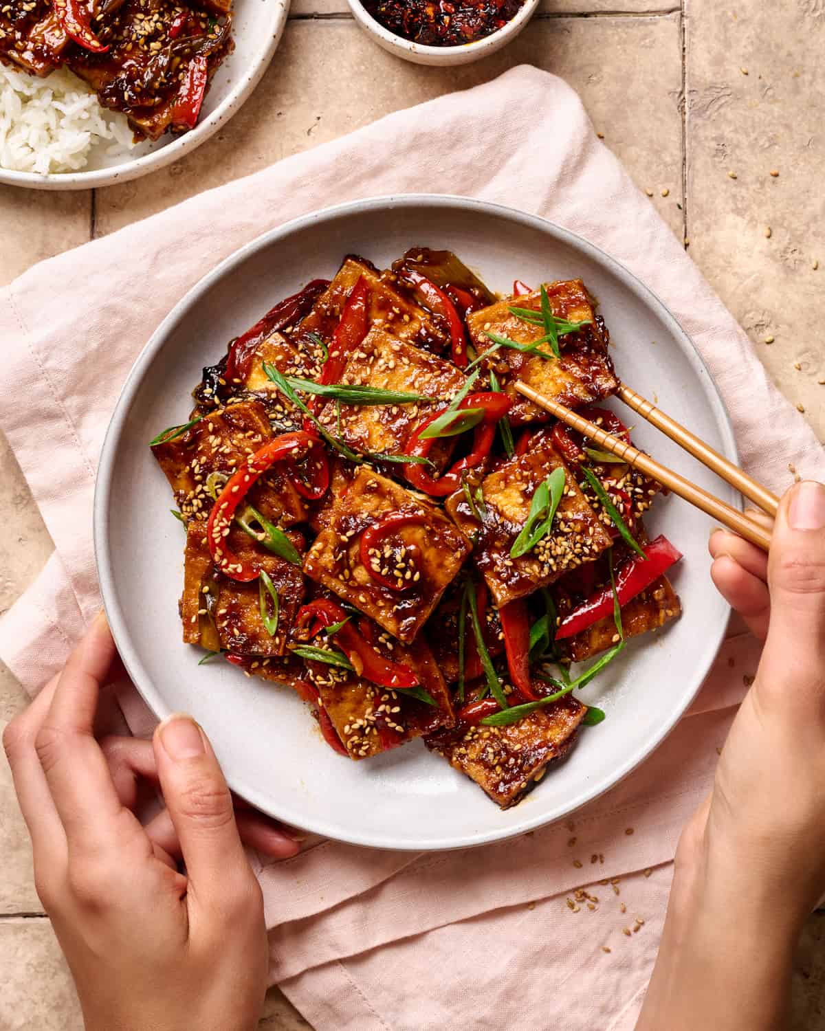 Person grabbing a piece of braised tofu from a white plate on a cream colored table.