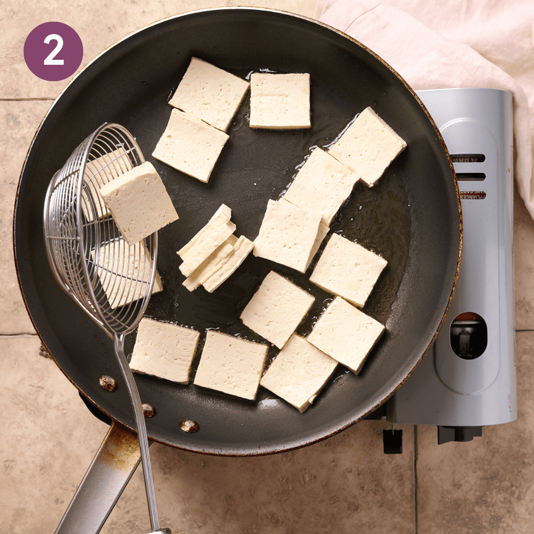 Person adding tofu to oil in a frying pan with a spider utensil.