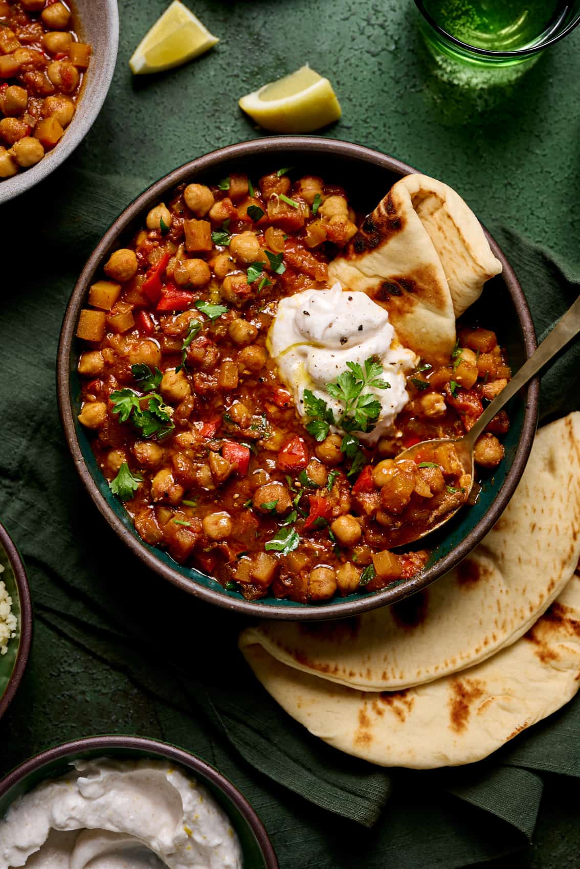 bowl of vegan tagine with chickpeas topped with yogurt and pita on a green table with green glasses.