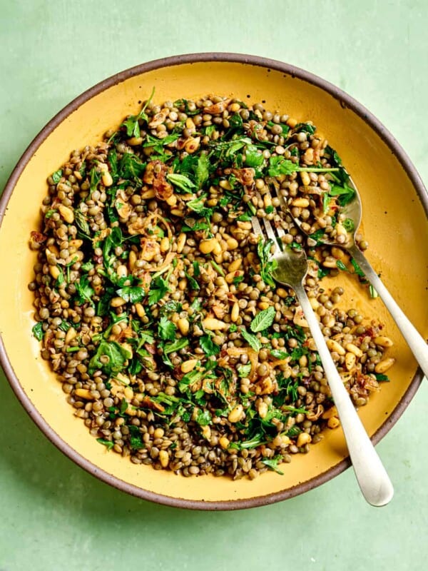 Spoon and fork in lentil salad in a yellow bowl on green table.
