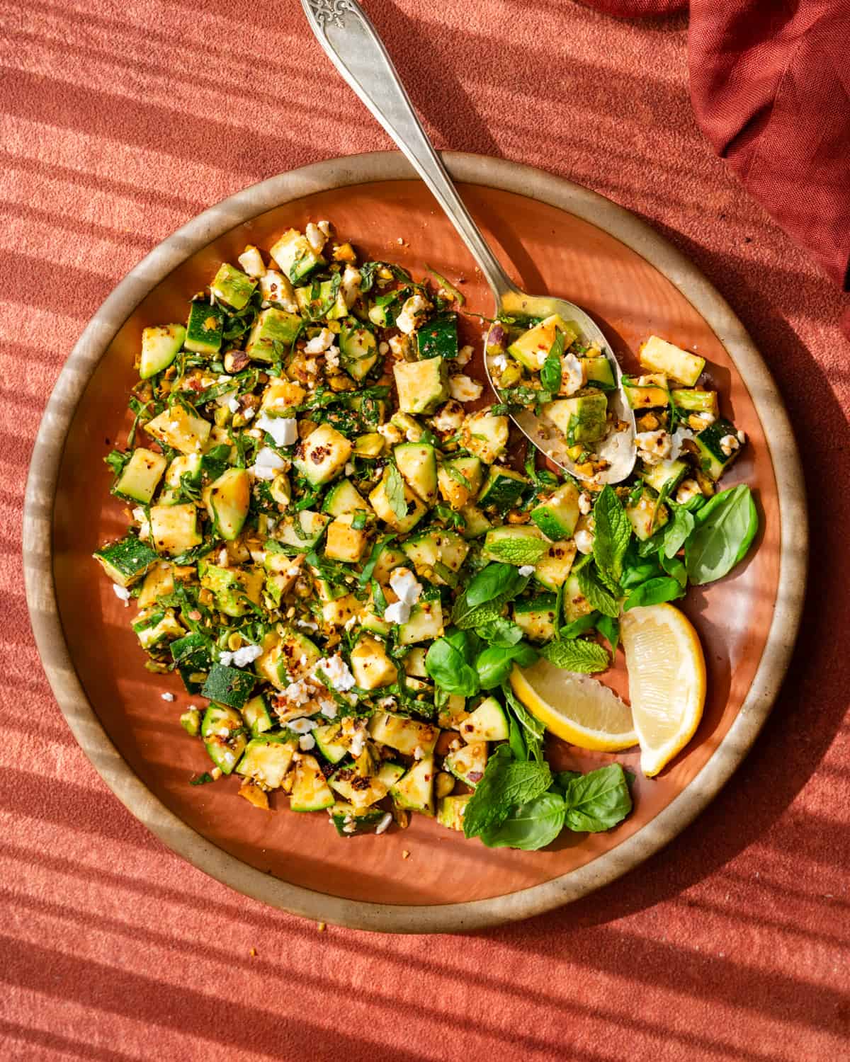 zucchini salad tossed with chili oil, pistachios, feta, basil, and mint on a red place on a red surface.