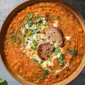 Bowl of red lentil soup soup with bread in it topped with yogurt and fresh herbs.