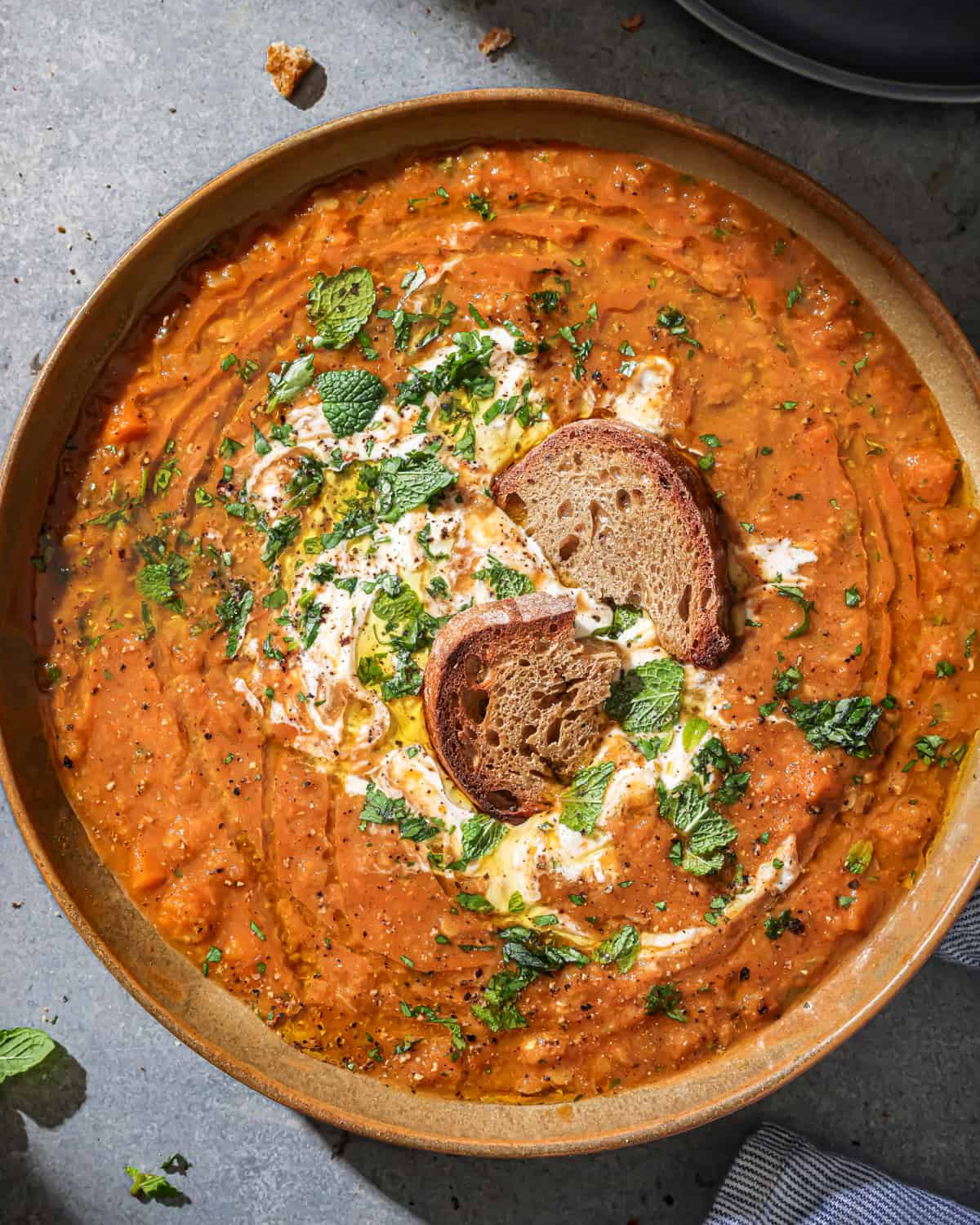 Bowl of red lentil soup soup with bread in it topped with yogurt and fresh herbs.